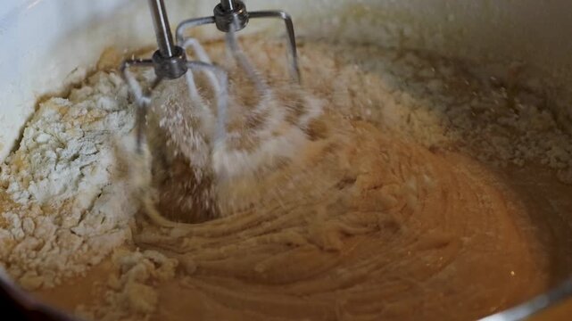 Closeup of rotating electric mixer beaters gradually incorporating dry flour into thick brown batter in a mixing bowl. 
