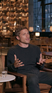Interview Session: A man in a black shirt is animatedly gesturing with his hands while engaged in a conversation, seated in a modern, softly lit lounge area with blurred background.