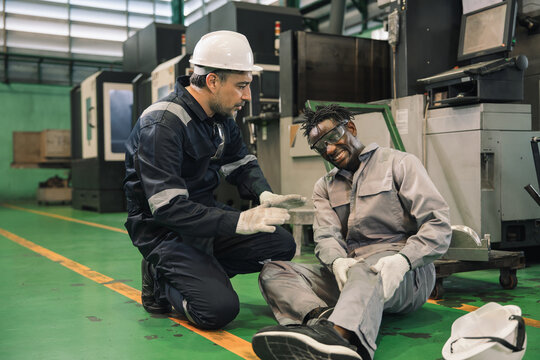 African engineer with hurting knee sitting on the floor and Caucasian worker in safety helmet helping and calling ambulance