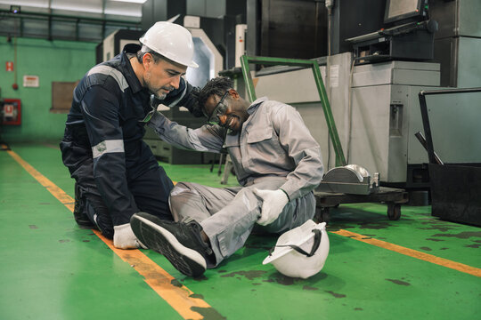 African engineer with hurting knee sitting on the floor and Caucasian worker in safety helmet helping and calling ambulance