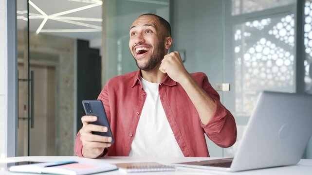 Happy excited businessman celebrates success by reading great news on mobile phone sitting at workplace in modern business office. Smiling shocked surprised entrepreneur received positive good message