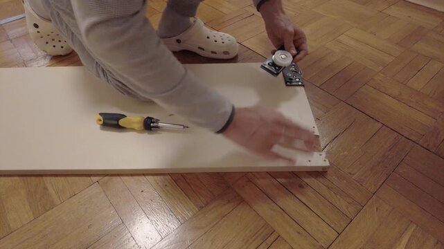 Closeup hands installing roller on cabinet homeowner kneels on parquet floor, inspects metal bracket and wheel against recessed hole, screwdriver and screws nearby, careful