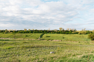 Fototapeta premium Wide panoramic view of a green meadow with rolling hills, trees, and dramatic clouds in the sky. Peaceful rural landscape illuminated by soft evening sunlight, showcasing natural beauty, open