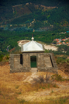 High angle view of a historic stone chapel on a mountain slope overlooking a vast green valley and village in Mondim de Basto, Portugal.
