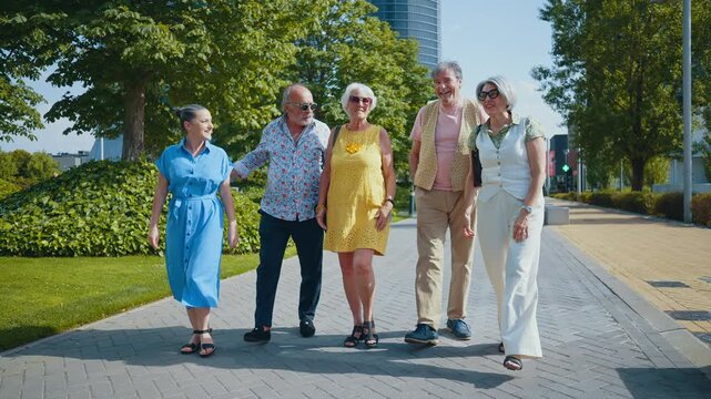 Group of five cheerful senior friends enjoying a sunny day, walking together along a paved path in a modern city park, laughing and talking with one another in a vibrant, friendly atmosphere