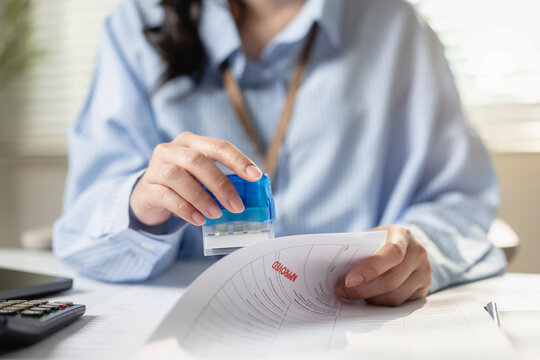 Female accountant stamping rubber stamp approved text on official contract document at desk in office. Concept of business approval process, agreement clearance, and official contract authorization.