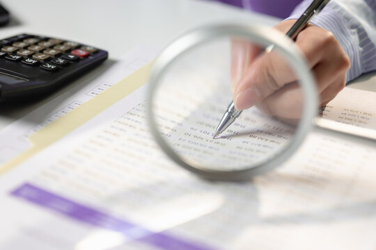 Close up hands of auditor holding pen inspecting financial report document paper through magnifying glass at desk. Concept of tax audit process, budget examination, and accounting analysis.
