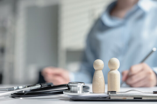 Close up wooden dolls life partner and stethoscope on clipboard with female doctor writing document in background. Concept of family health insurance, medical coverage, and healthcare protection plan