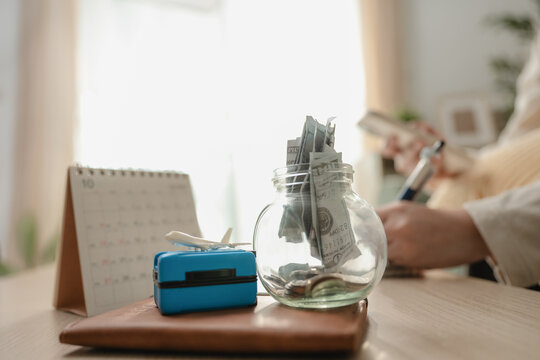 Close up glass jar filled with money cash savings, miniature luggage suitcase, toy airplane, and desk calendar on table. Concept of travel budget savings, vacation financial planning, and future trip.