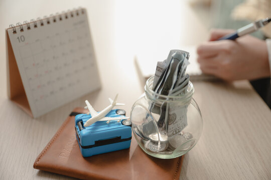 Close up glass jar filled with money cash savings, miniature luggage suitcase, toy airplane, and desk calendar on table. Concept of travel budget savings, vacation financial planning, and future trip.