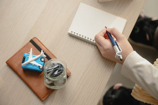 Top view hand writing travel budget on blank notebook desk near passport, savings money jar, miniature suitcase, airplane model. Concept of trip planning and vacation financial preparation.