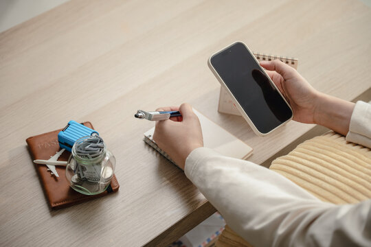 Woman holding mobile phone with blank screen writing notebook travel budget planning near money savings jar desk calendar. Concept of vacation financial planning, trip calculation, and online app.