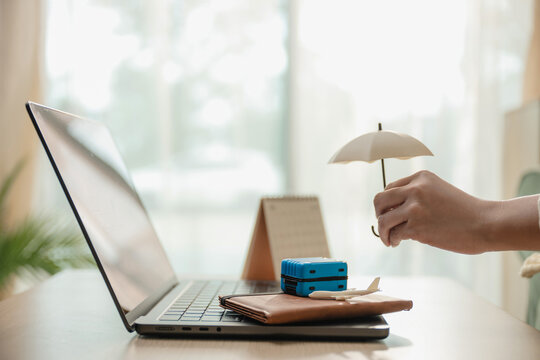 Hand holding small umbrella protecting miniature luggage, toy airplane, passport on laptop desk. Concept of travel insurance coverage, flight booking protection, and vacation safety with copy space.