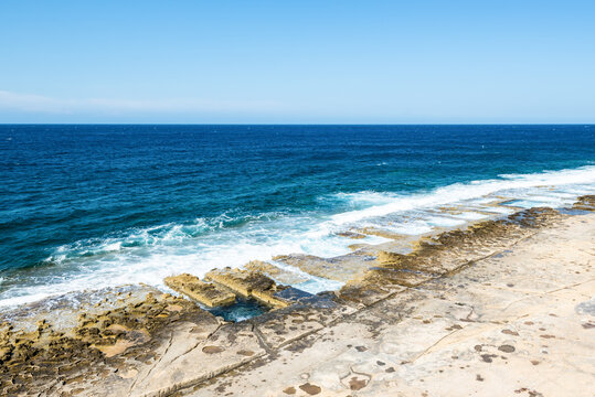 Scenic view of the historic man-made Roman baths carved into Sliema&rsquo;s rocky coastline, with turquoise Mediterranean waters and waves washing over the limestone shore in Malta
