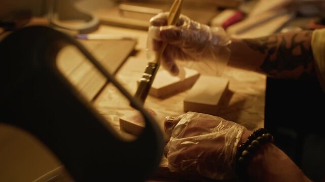 Caucasian male artisan wearing gloves dipping paintbrush into cup and staining wooden blocks at workbench. Footage is perfect for DIY craft tutorials and small business marketing campaigns