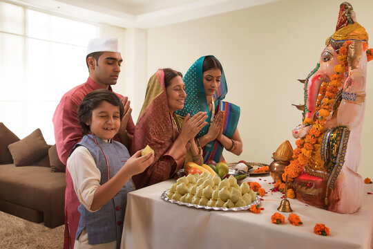 Family praying on Ganesh Chaturthi folding hands where the child winks and takes a modak from the prasad