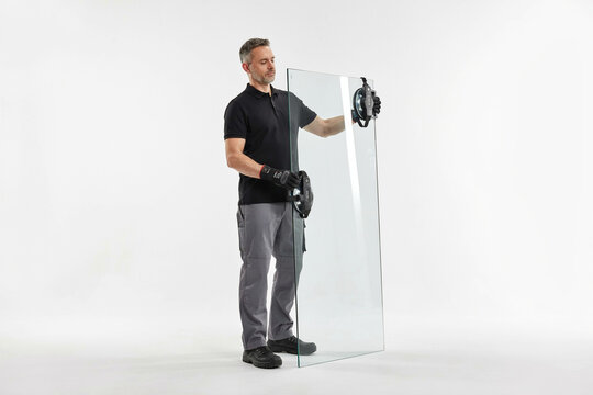 Professional glazier carrying large clear glass panel with suction lifter, construction worker in black shirt and gloves isolated on white background