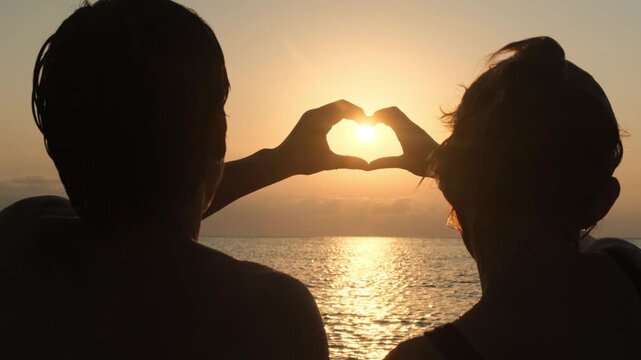 Silhouettes of a young couple making a heart with joined hands at sunset over the sea horizon. Love will save the world. Romance, loyalty and lasting relationships.
