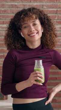 Video of sporty woman with detox juice looking at camera while listening music with earphones in the kitchen at home.