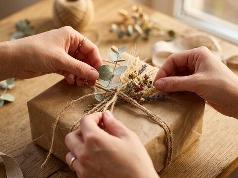 Macro view of hands carefully decorating a brown paper parcel with dried botanical elements and twine during the warm golden hour sunlight.