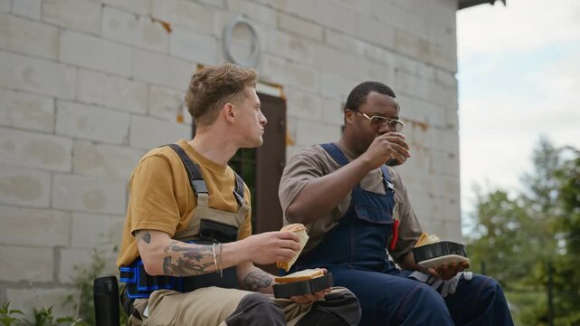 Caucasian and African American young adult builders sitting outside unfinished house, eating lunch and talking. Video is ideal for construction industry marketing campaigns