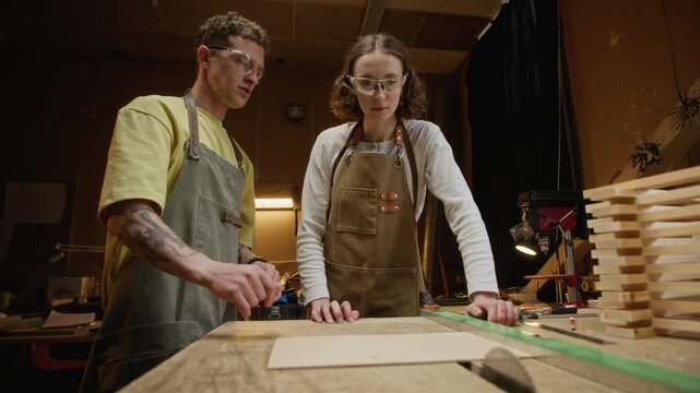 Young adult Caucasian man and woman working in carpentry workshop, discussing wood cutting process on table saw. Clip is perfect for vocational school and craft business promotions