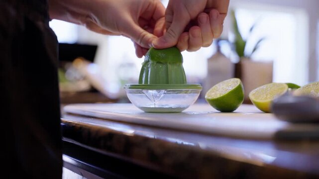 Close-up of a hand pressing a lime in a green manual citrus juicer, with halved limes on a kitchen counter and bright home cooking atmosphere.