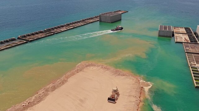 Aerial view of a large bulldozer pushing sand into the turquoise sea, contributing to a land reclamation project for coastal development while a tugboat navigates nearby concrete caissons
