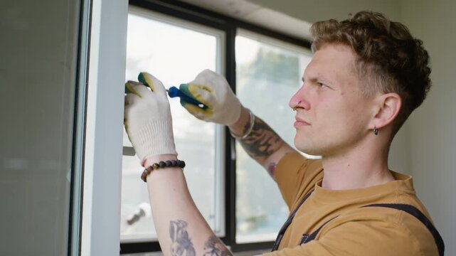 Young adult Caucasian male handyman wearing gloves installing window frame with screwdriver during home renovation. Video is perfect for construction services and DIY home improvement marketing