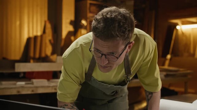 Caucasian young adult male carpenter measuring wood with ruler and pencil on blueprint in workshop. Footage is suitable for promoting small businesses, craftsmanship, and vocational education