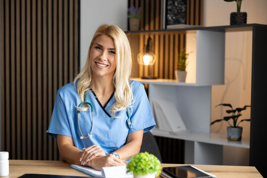 Confident female doctor wearing blue scrubs and stethoscope, sitting at a desk in a modern medical clinic, smiling