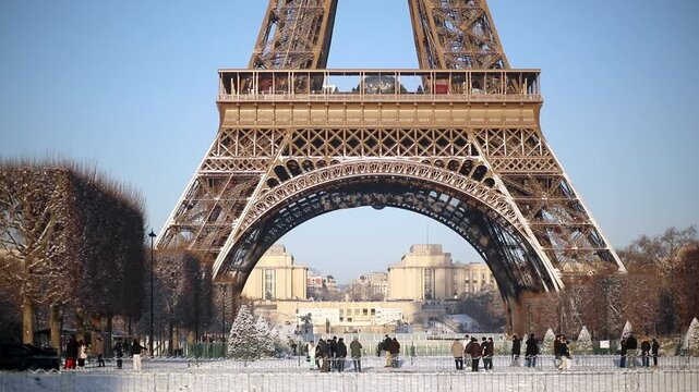 Wide view of the Eiffel Tower with its base surrounded by snow-covered trees and park grounds in Paris. Captured on a bright winter morning, with people walking in the distance and long shadows.