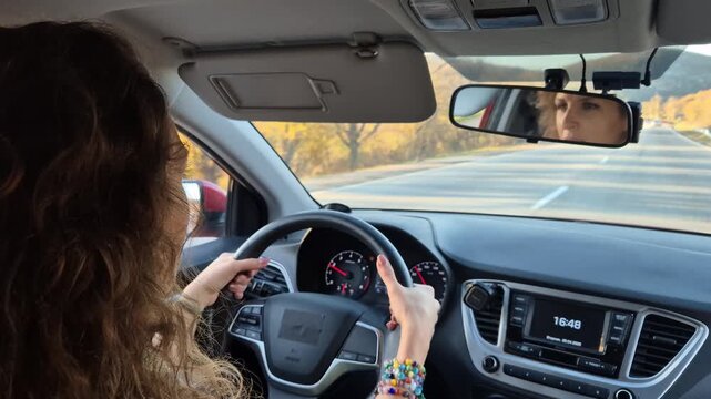 Woman driving car along scenic highway during sunny golden hour while holding steering wheel with colorful bracelets. Interior static back seat shot.