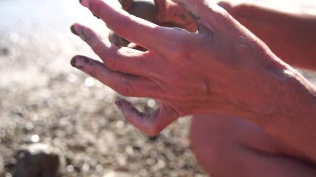 Hands mud creativity, person playing with wet clay on a sunny pebble beach, forming a small round ball, childhood memory nature.