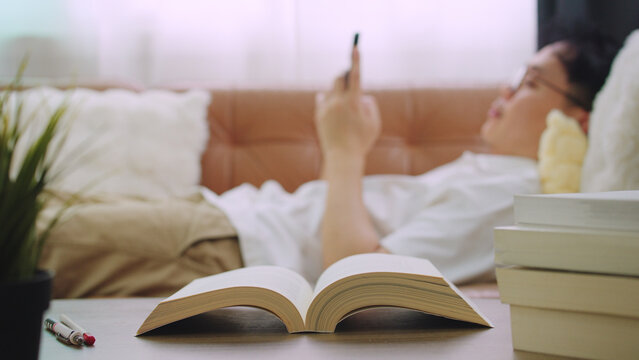 Young Man Lying on Sofa Browsing Smartphone with Open Book in Foreground Focus.