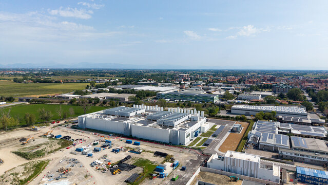 Aerial view of Vantage Data Centre - MXP1, a large industrial facility with white roofs and cooling units, surrounded by green fields and urban infrastructure in Melegnano, Lombardia, Italy.