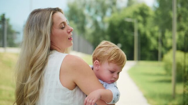 Mother carries baby outdoors on sunny day. Baby looks around curiously. Mother smiles gently, enjoying moment. Ideal for family, parenting, or nature-themed projects