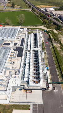 Aerial view of Vantage Data Centre - MXP1 with rows of industrial cooling units and rooftop infrastructure, adjacent to railway tracks and green fields in Melegnano, Lombardia, Italy.