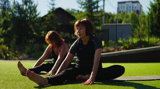 Elderly women practicing gentle Pilates stretching outdoors on grass, performing slow motion seated flexibility exercises for senior fitness and healthy aging