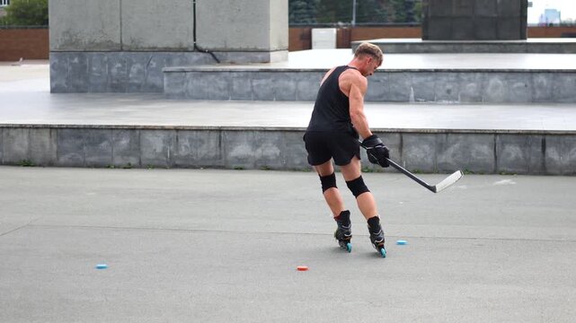 Slow motion video of a middle-aged man practices hockey stick techniques while roller skating on an asphalt rink in the summer. Dolly shot 4k footage.