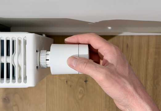 Hand adjusts a thermostatic radiator valve on a white heater. Close up of home heating control in an indoor interior. Energy saving temperature regulation for winter comfort..