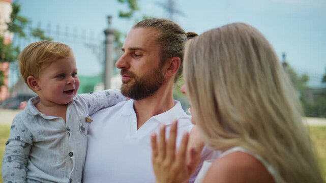 Young father holds smiling toddler while mother laughs nearby. Warm outdoor moment captures family joy and connection. Ideal for parenting, family, or lifestyle content