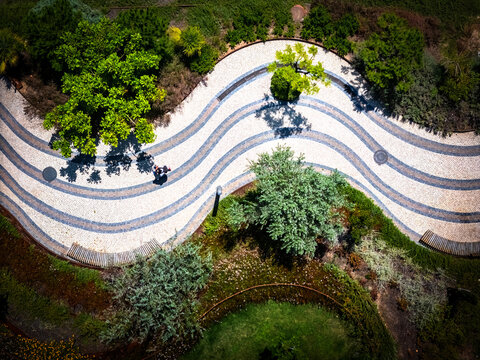 Aerial view of a winding cobblestone path with wavy patterns surrounded by lush green trees and manicured gardens in Cascais, Porto, Portugal.