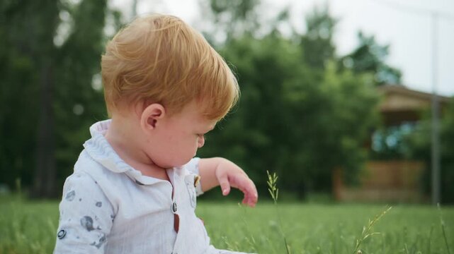 Young child sits in grass, eyes closed, hand reaching out. Soft light bathes the scene. He appears calm and curious. Perfect for nature, childhood, or peaceful moments. Can be used in advertising