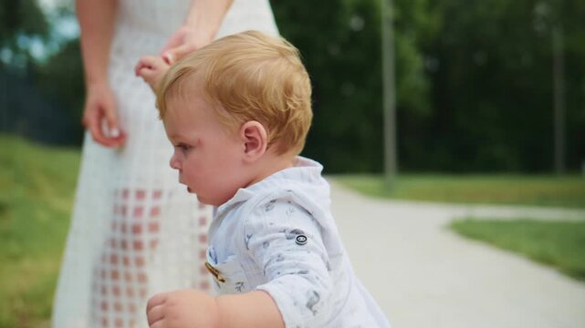 Toddler holds adult's hand while walking outdoors. Child looks upward with curious expression. Background shows green park pathway. Footage captures tender moment between caregiver and child