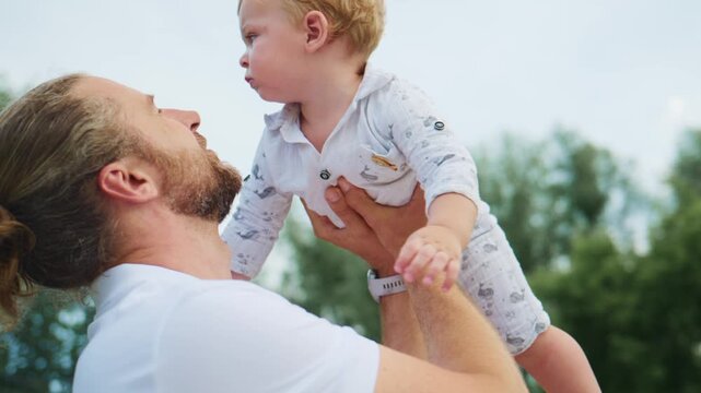 Man lifts baby up outdoors, baby looks upward with curiosity. Mother holds baby from below, both smile gently. Scene captures playful, loving family interaction. Ideal for parenting, childhood