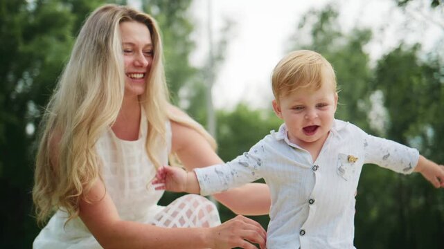 Mother laughs while holding toddler outdoors. Child smiles broadly, arms outstretched. Green trees softly blur background. Joyful family moment captures natural playtime. Perfect for parenting