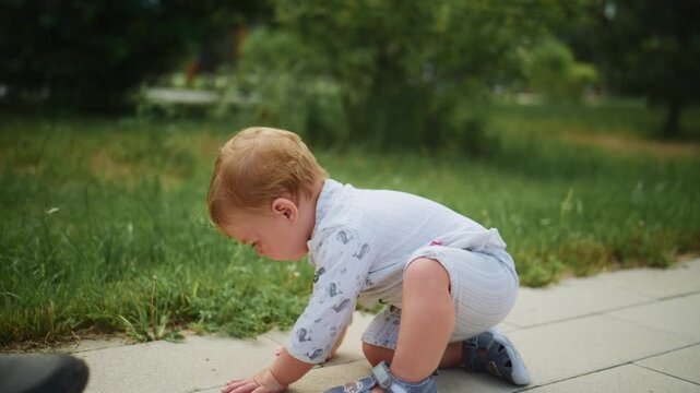 Young child crawling on path, smiling joyfully. Hand gently supports toddler's back. Outdoor park scene with green grass and trees. Ideal for family, growth, or childhood development themes