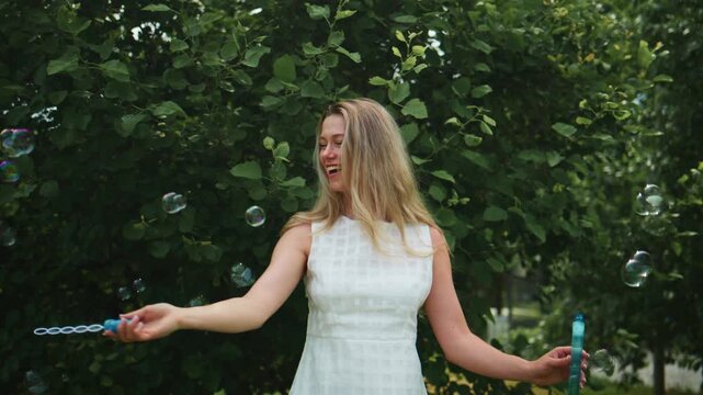 Woman in a white dress blows soap bubbles outdoors. She smiles joyfully as bubbles float around her. Green trees frame the scene with soft, natural lighting. Ideal for advertising summer fun