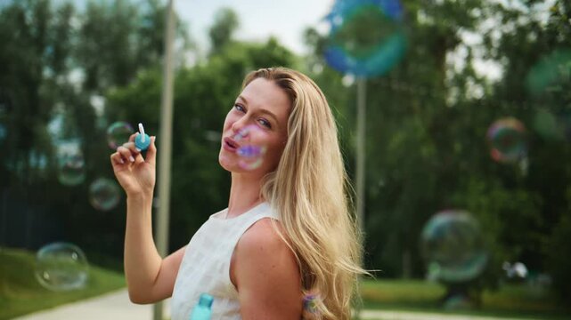 Woman blows soap bubbles in sunny park. She smiles and enjoys summer day. Peaceful outdoor fun for advertising or lifestyle content. Gentle wind carries floating bubbles. Perfect for vacations